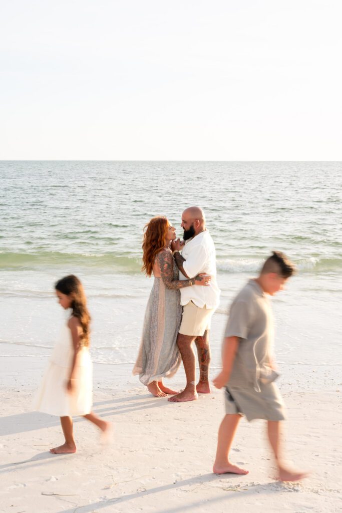Parents embracing on beach while children walk nearby during Bean Point Beach family photography session on Anna Maria Island