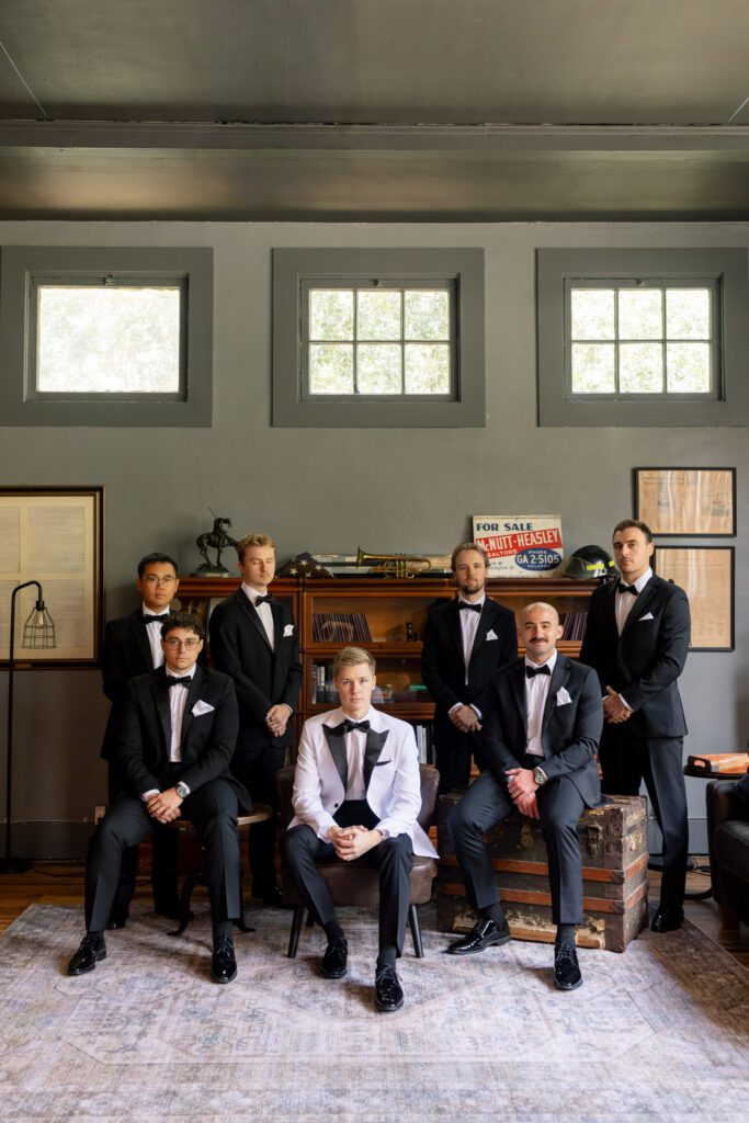 Groom and groomsmen in classic black tuxedos posing in a styled interior space with vintage decor and natural light, photographed by a Central Florida wedding photographer