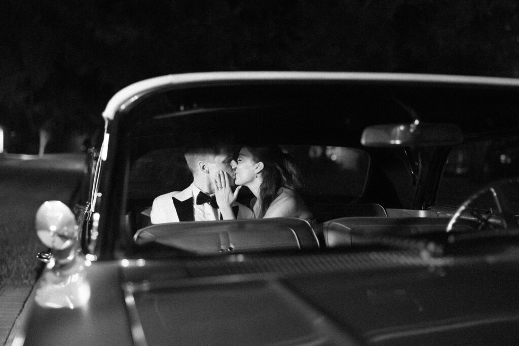 Black and white image of the couple sharing a kiss inside a vintage car, capturing a timeless and romantic moment during a Historic Venue 1902 wedding