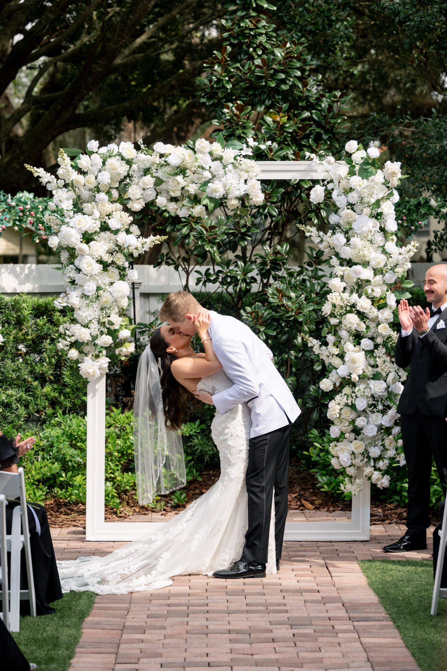 Couple sharing their first kiss beneath a white floral ceremony arch surrounded by greenery during a romantic Sanford Florida wedding