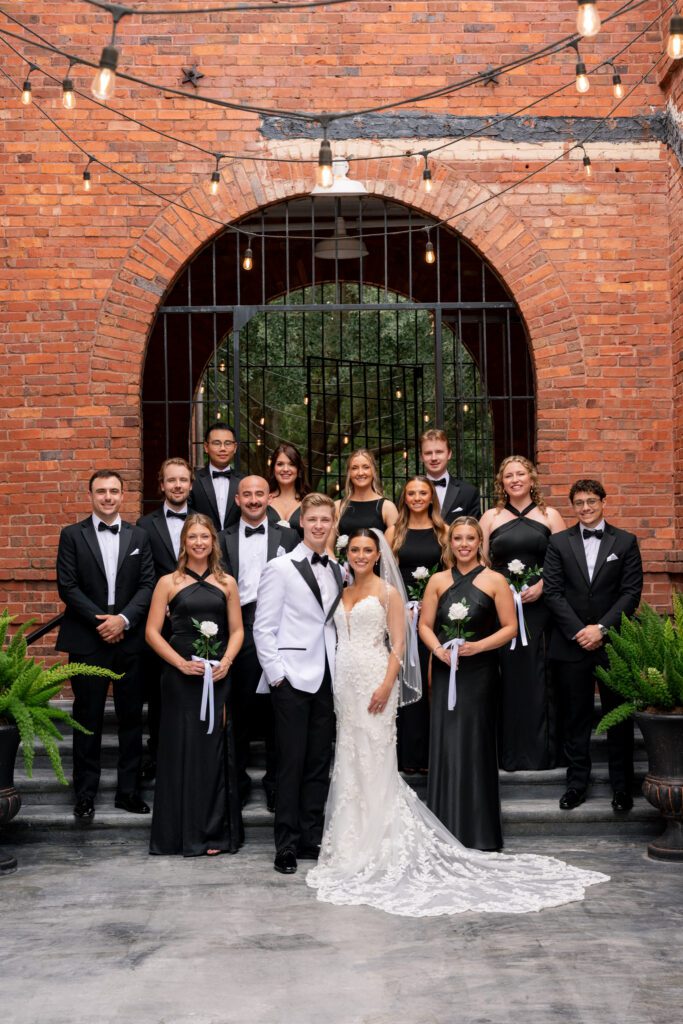 Bride and groom posing with their wedding party in front of a historic brick building with string lights and arched entryway at a Historic Venue 1902 Sanford wedding