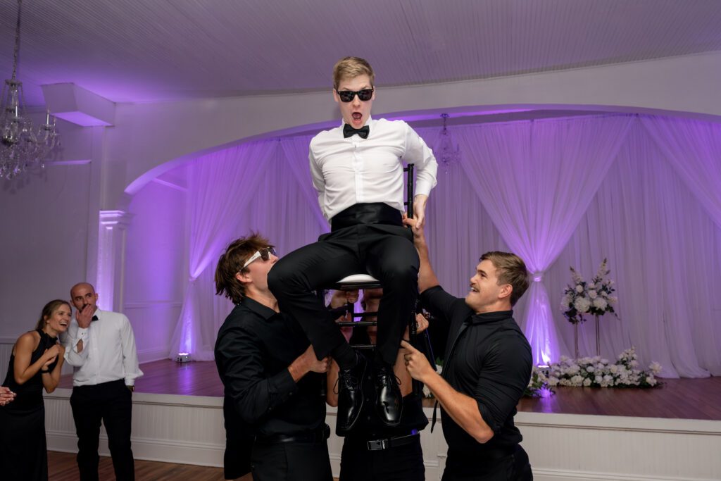 Groom being lifted on a chair by friends during a lively reception celebration with music and dancing at a Historic Venue 1902 Sanford FL wedding