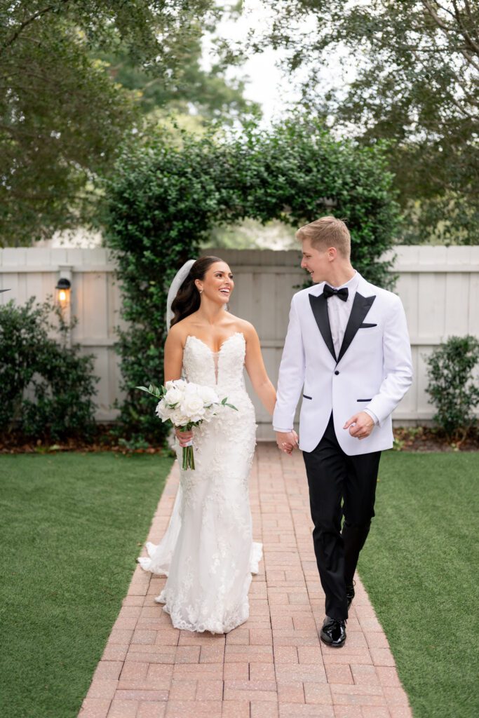 Bride and groom walking hand in hand along a garden pathway, smiling at one another in a classic outdoor portrait at a Historic Venue 1902 Sanford FL wedding