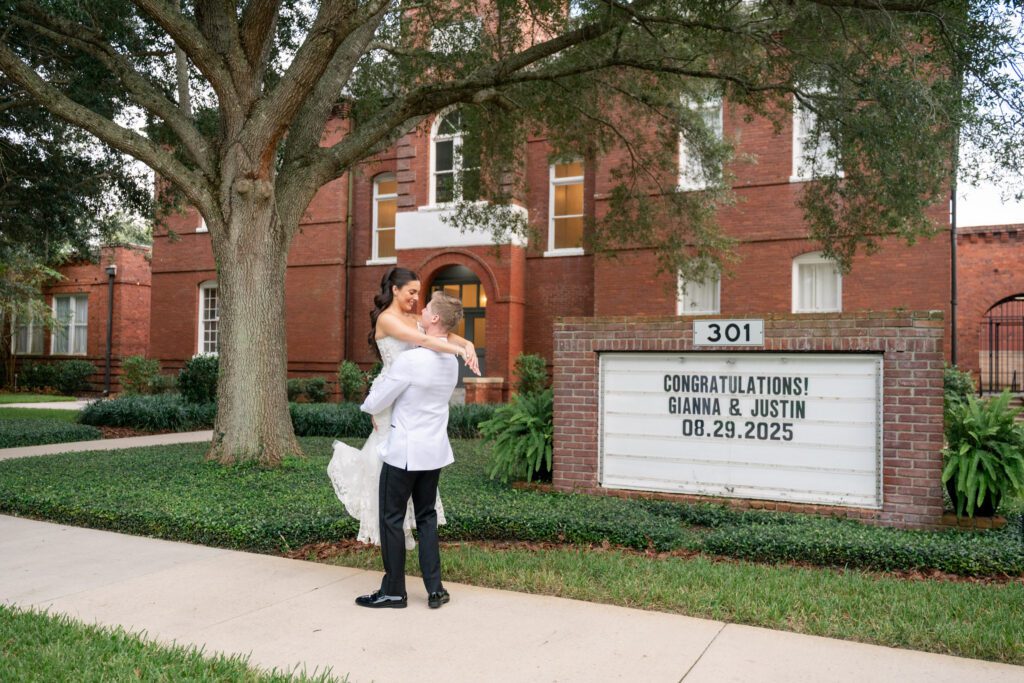 Groom lifting the bride in front of a historic brick building and marquee sign displaying their wedding date at a Historic Venue 1902 Sanford wedding