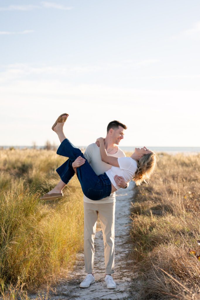 Playful couple portrait with groom lifting bride along a sandy path during a Siesta Key engagement session near Sarasota’s Gulf Coast dunes