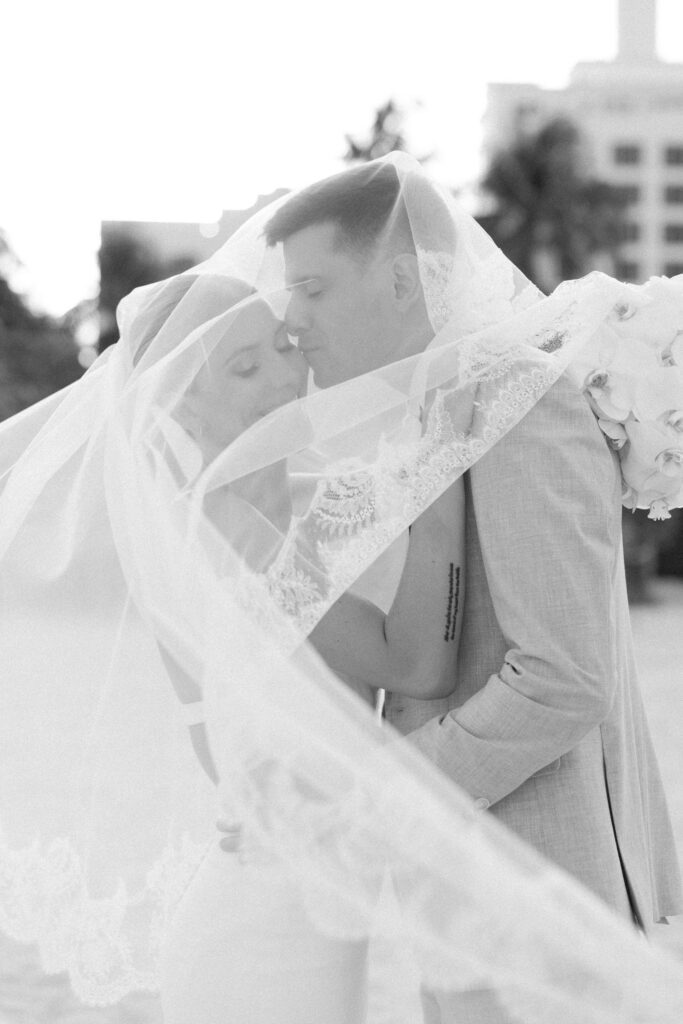 Black and white portrait of a newlywed couple embracing under a flowing veil, capturing an intimate moment during a Miami Beach destination wedding