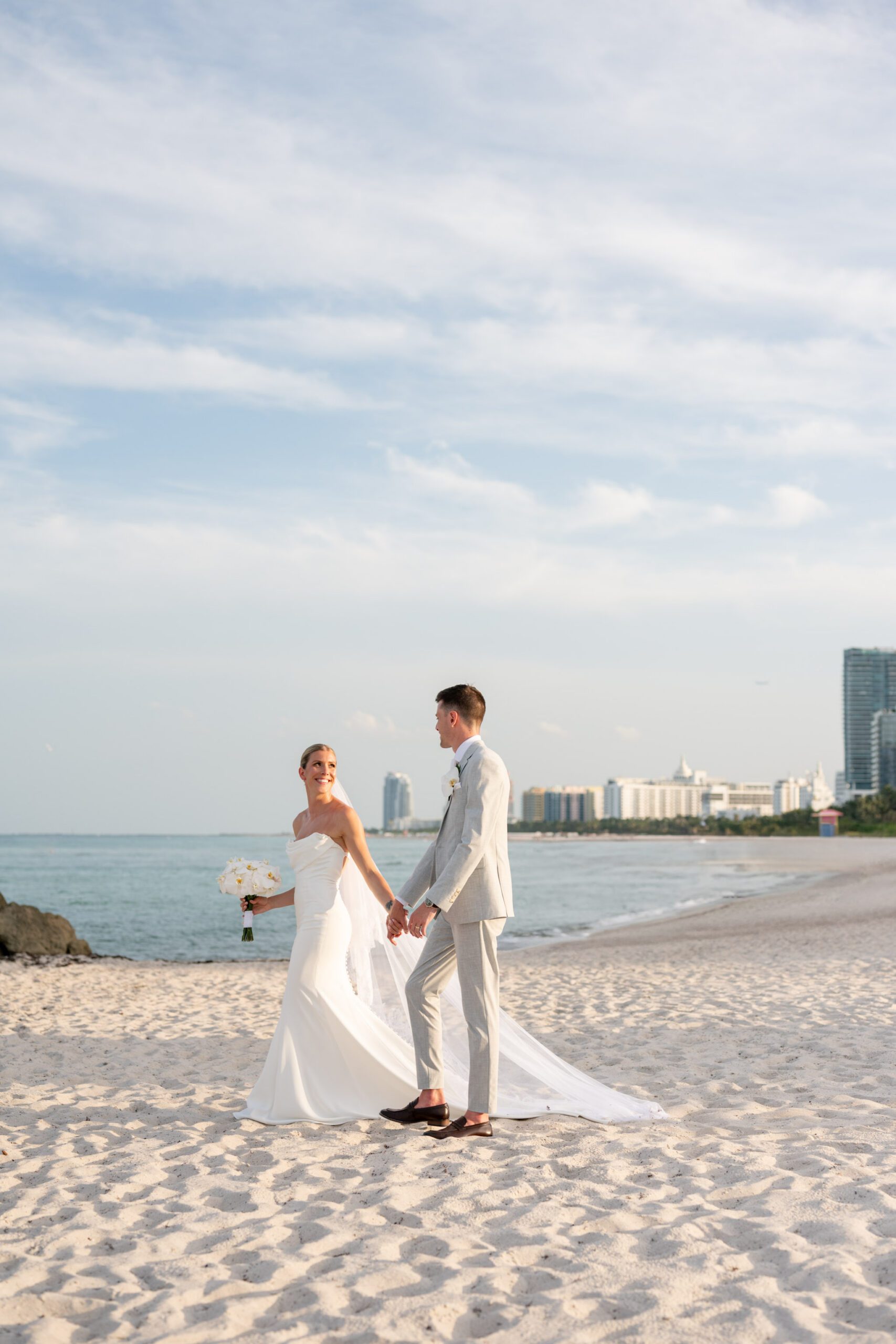Bride and groom walking hand in hand along a sandy beach with ocean views and a soft flowing veil, captured during a coastal ceremony at The Palms Hotel & Spa Miami Beach wedding
