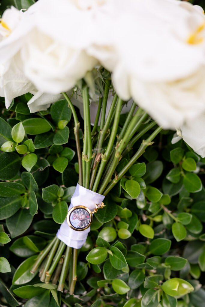 Close-up of a white rose bridal bouquet with greenery and a sentimental photo charm wrapped around the stems, captured in The Palms Hotel & Spa wedding photos