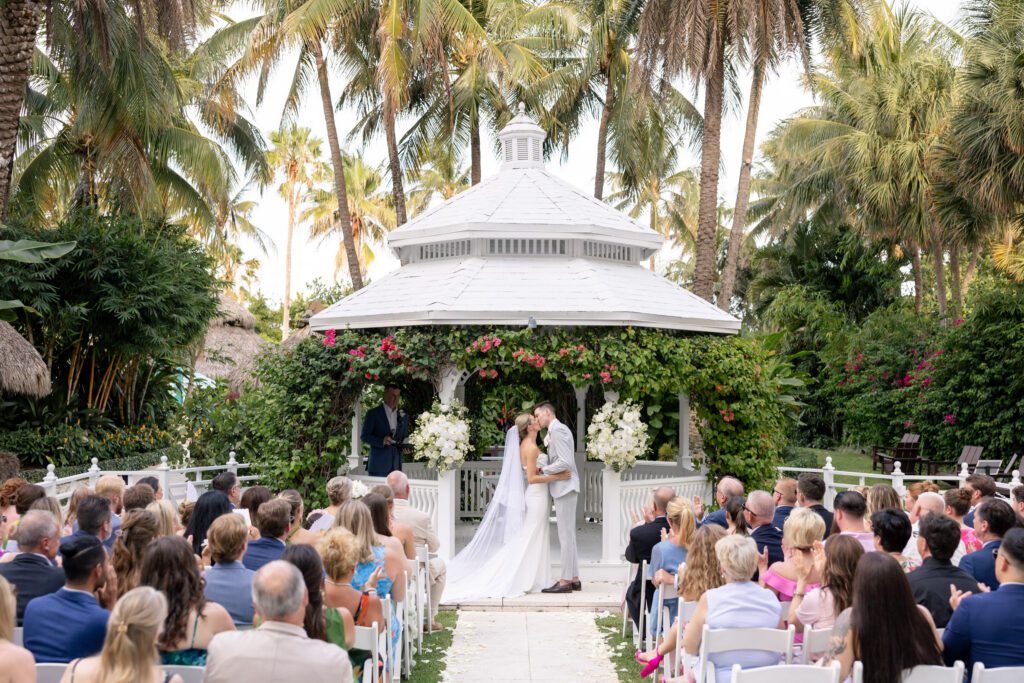 Bride and groom sharing a kiss at a white gazebo ceremony surrounded by lush greenery and guests during a tropical celebration at The Palms Hotel & Spa Miami wedding