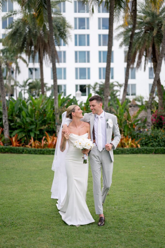 Newlywed couple smiling and walking across a lush green lawn surrounded by palm trees and tropical landscaping at The Palms Hotel Miami Beach wedding venue, photographed by a Miami Beach wedding photographer