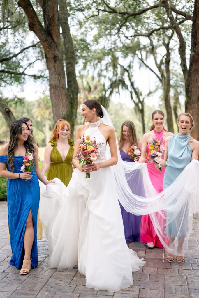 Bride walking with bridesmaids in colorful dresses while holding a flowing veil and vibrant bouquet during an outdoor Up The Creek Farms Melbourne FL wedding