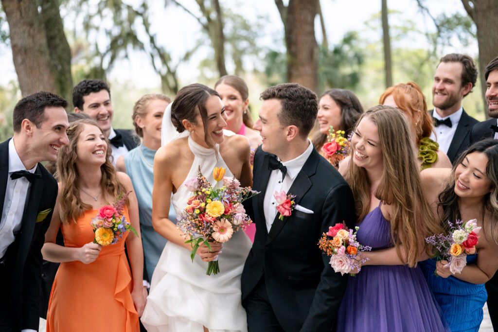 Bride and groom laughing together while walking with their wedding party and holding colorful florals during an outdoor Up The Creek Farms wedding